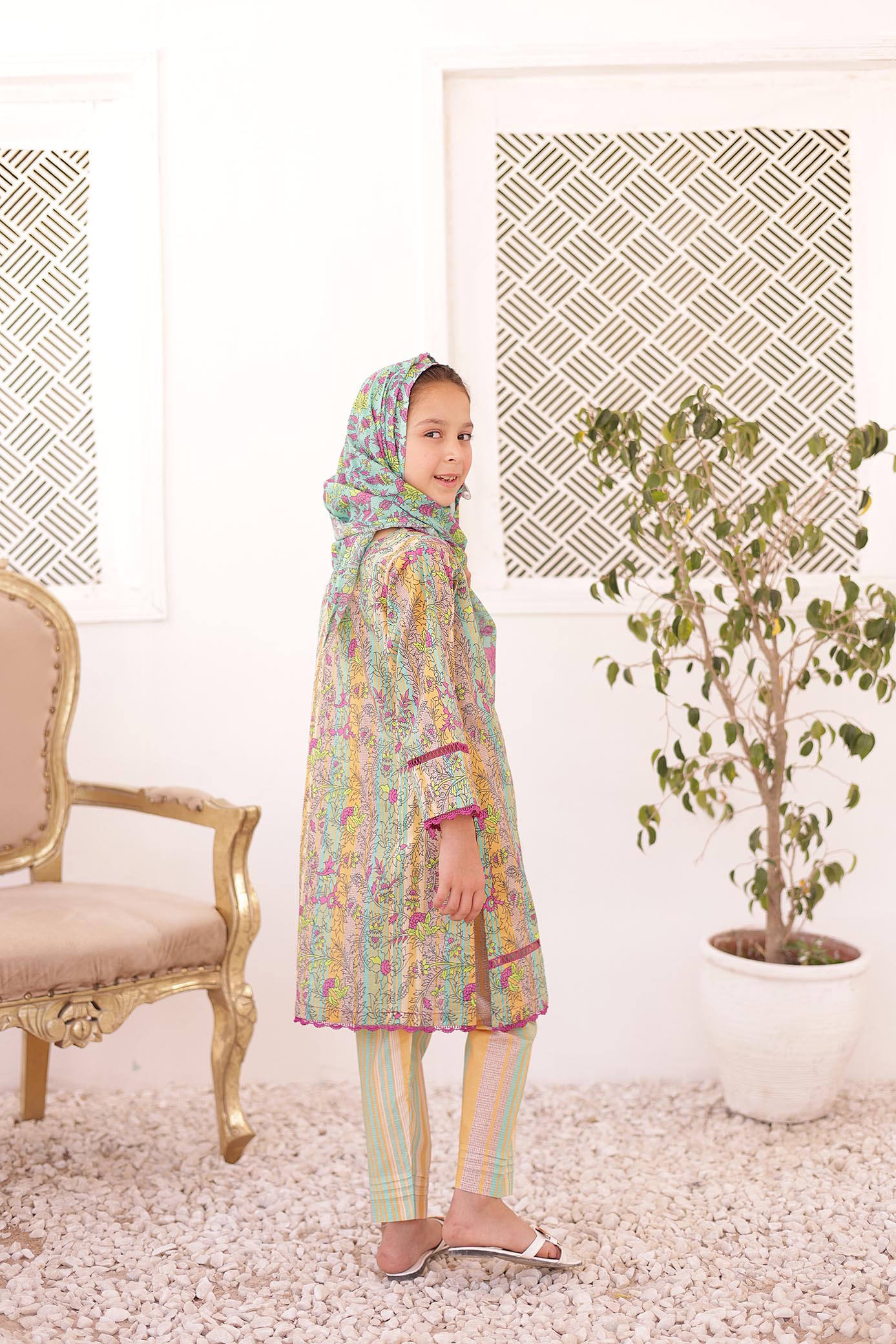 Child wearing a colorful traditional outfit in a room with a chair and plant.