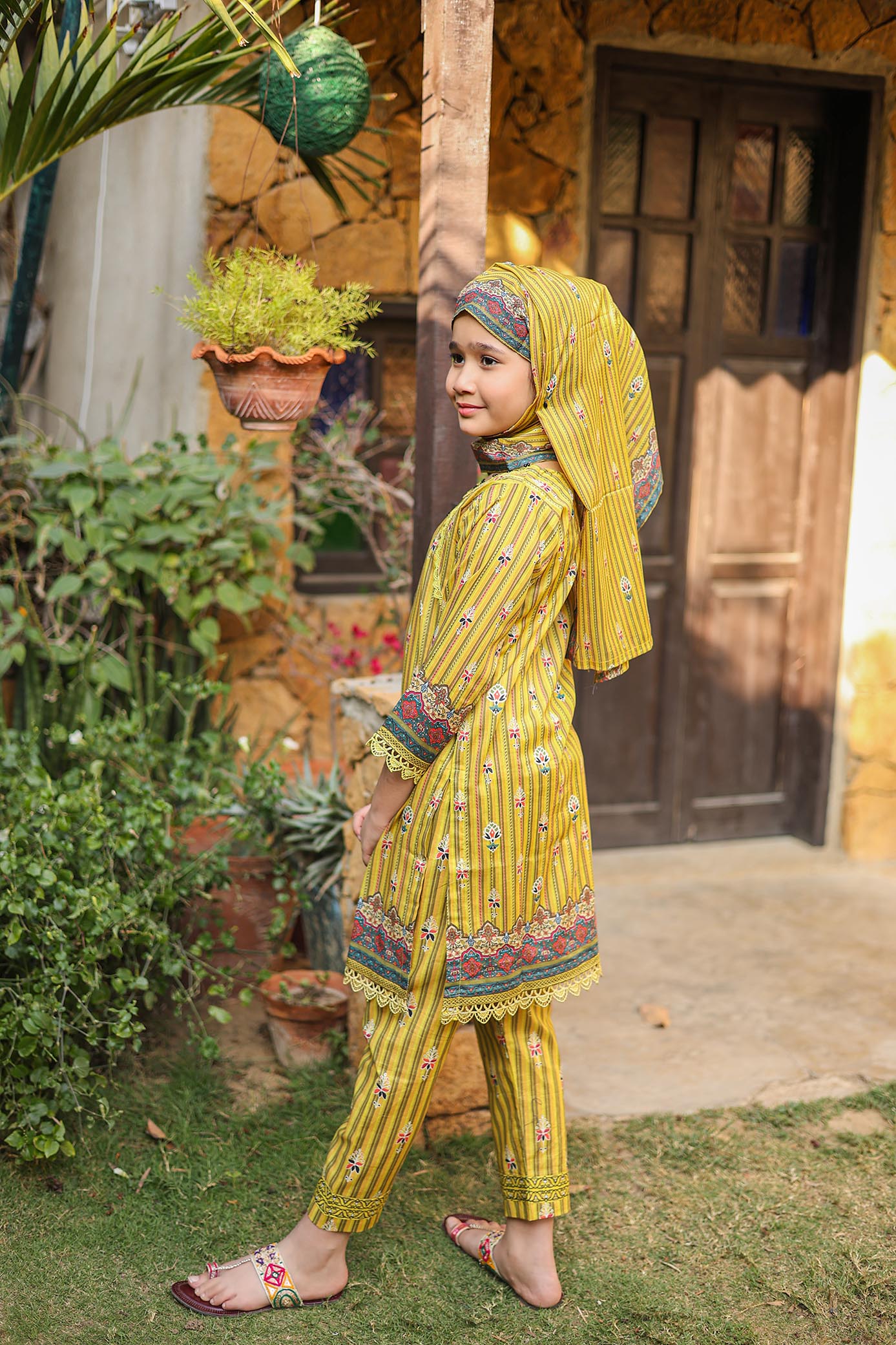 Child in a yellow traditional outfit standing outdoors with plants and a wooden door in the background