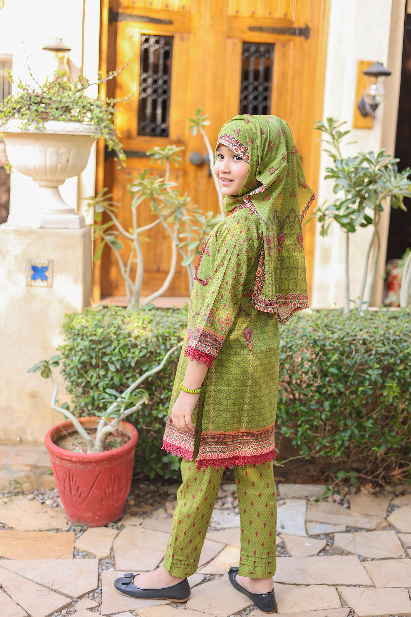 Woman in green traditional outfit standing outdoors with plants and a wooden door in the background