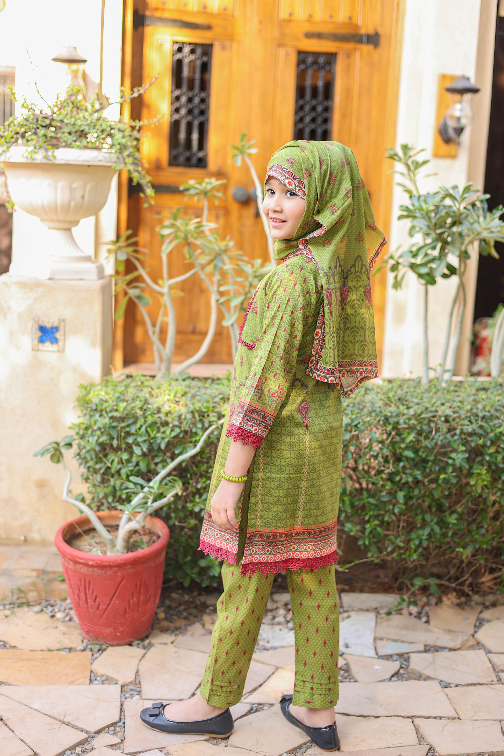 Woman in green traditional outfit standing outdoors with plants and a wooden door in the background