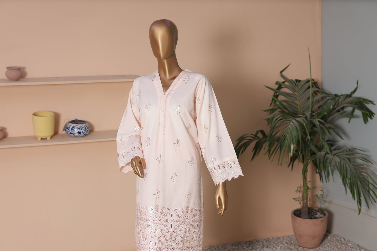 Mannequin wearing a white lace dress against a beige wall with decorative items and a plant.