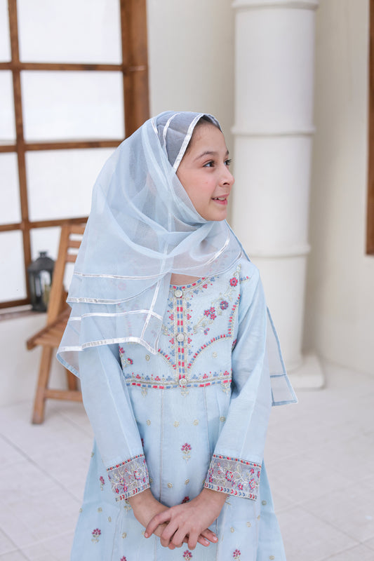 Young girl wearing a light blue embroidered dress and headscarf indoors.