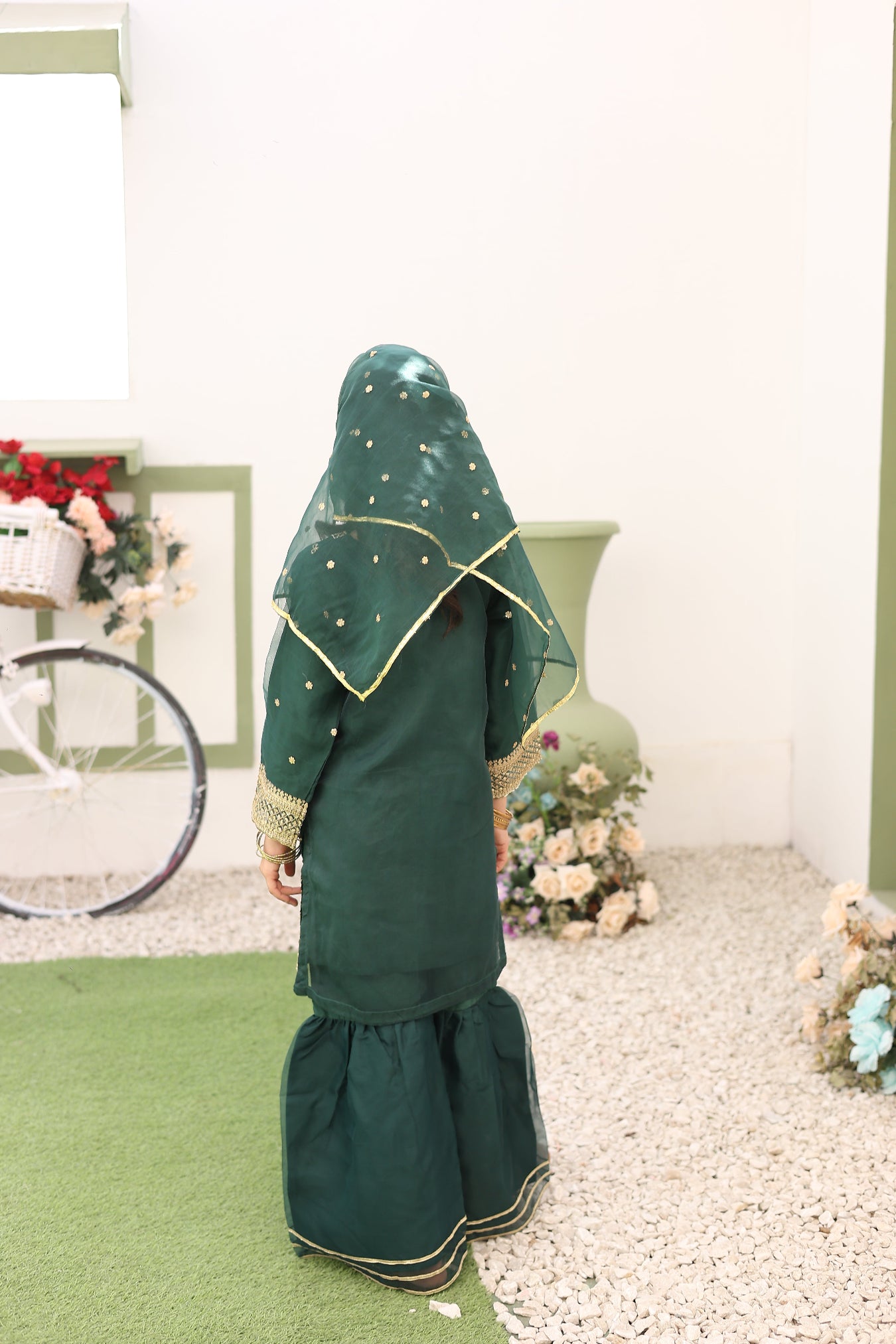 Person wearing a green dress with gold trim in a room with a bicycle and plants.