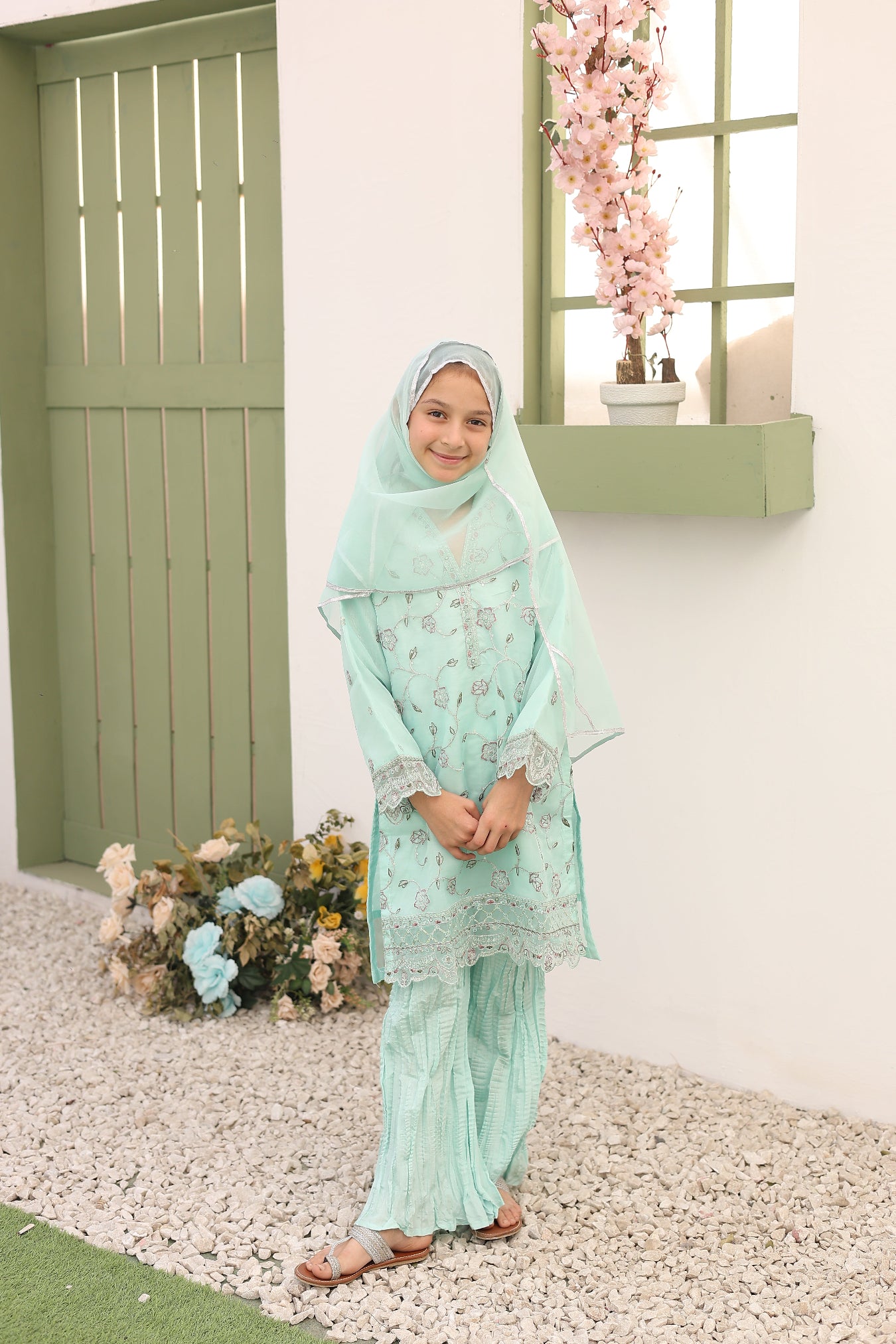 Woman in a light green dress with a matching headscarf standing in front of a green door with flowers.