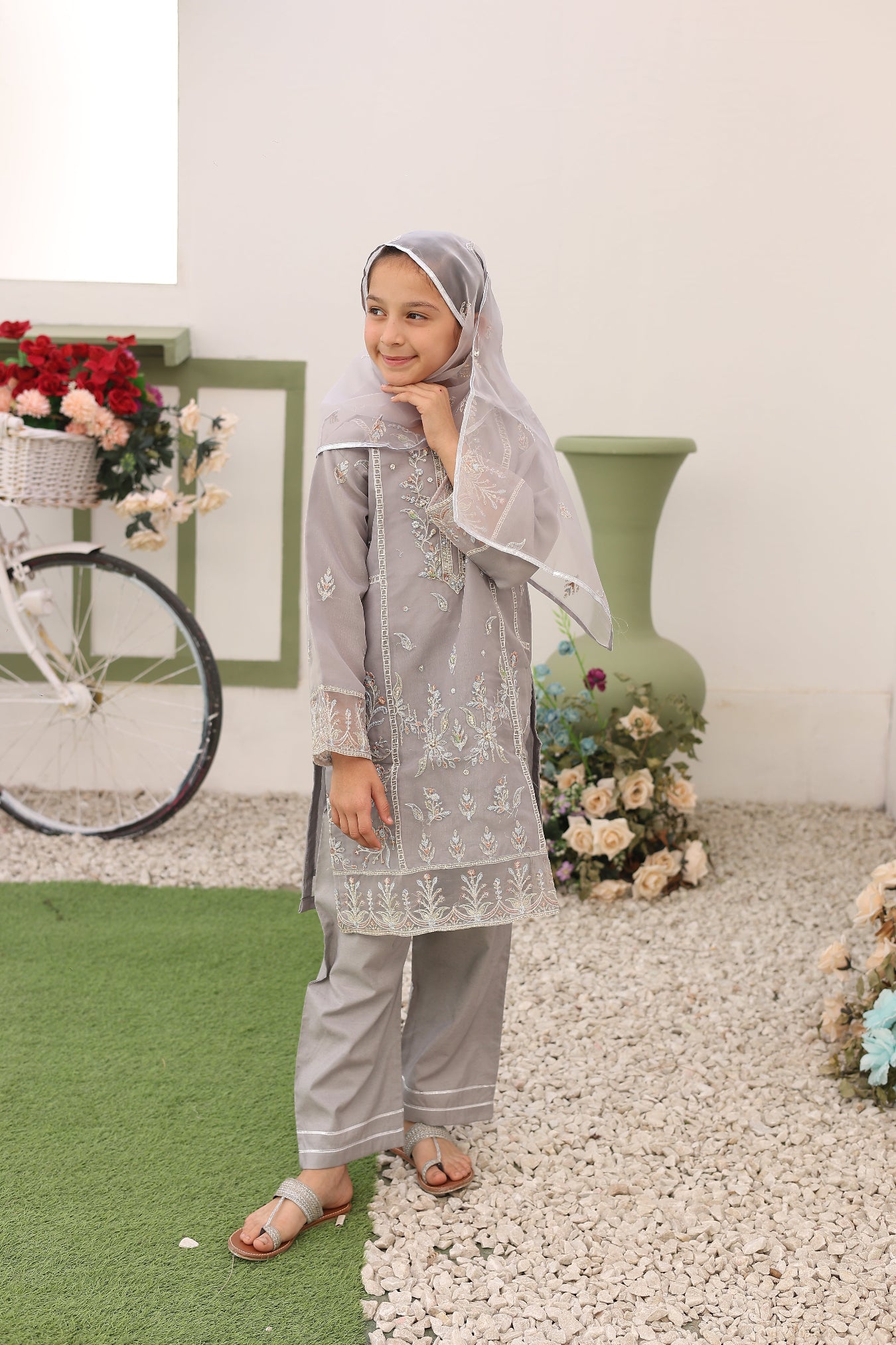 Woman in a gray traditional outfit standing on a checkered floor with flowers around her.