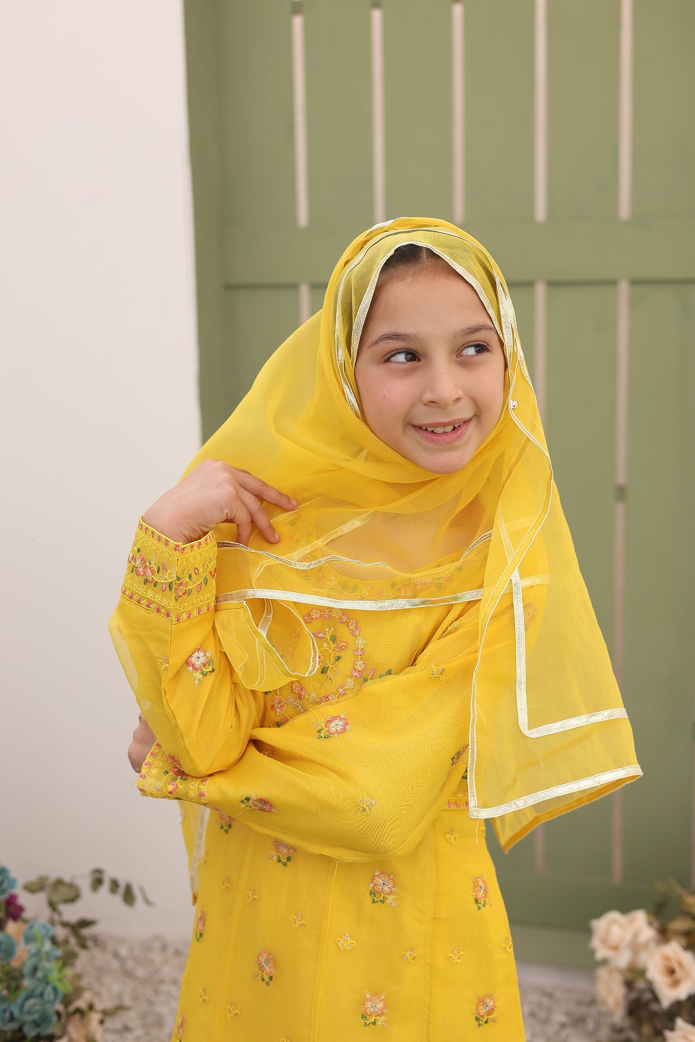 Child wearing a yellow embroidered outfit with a hijab, standing against a light-colored wall.