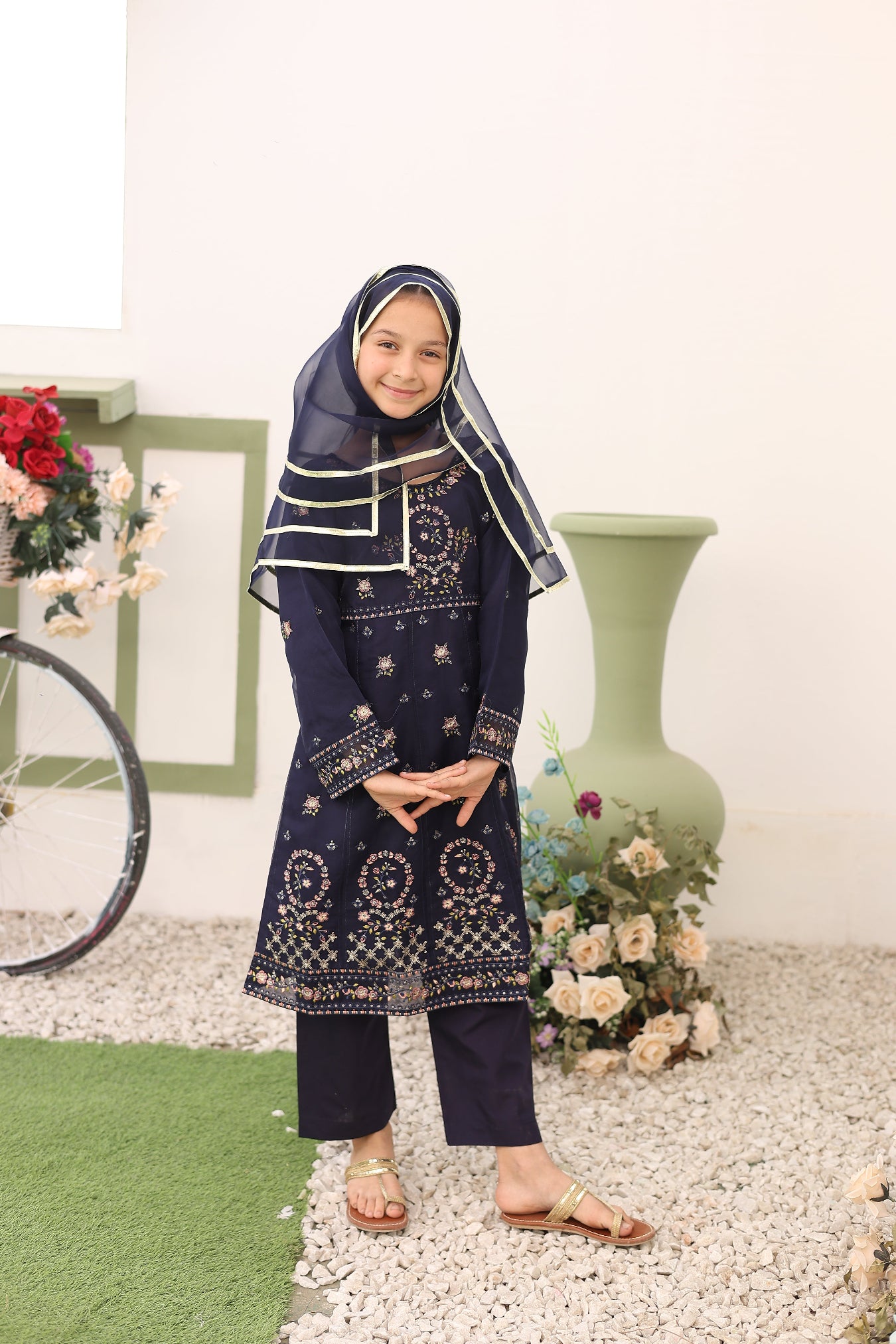 Woman in a navy blue traditional outfit with white patterns standing on a stone path with flowers and a green chair in the background.