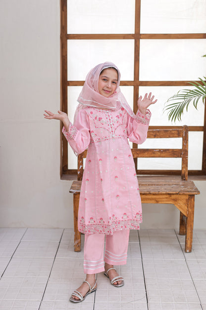 Woman in a pink traditional outfit standing in a room with wooden bench and window.