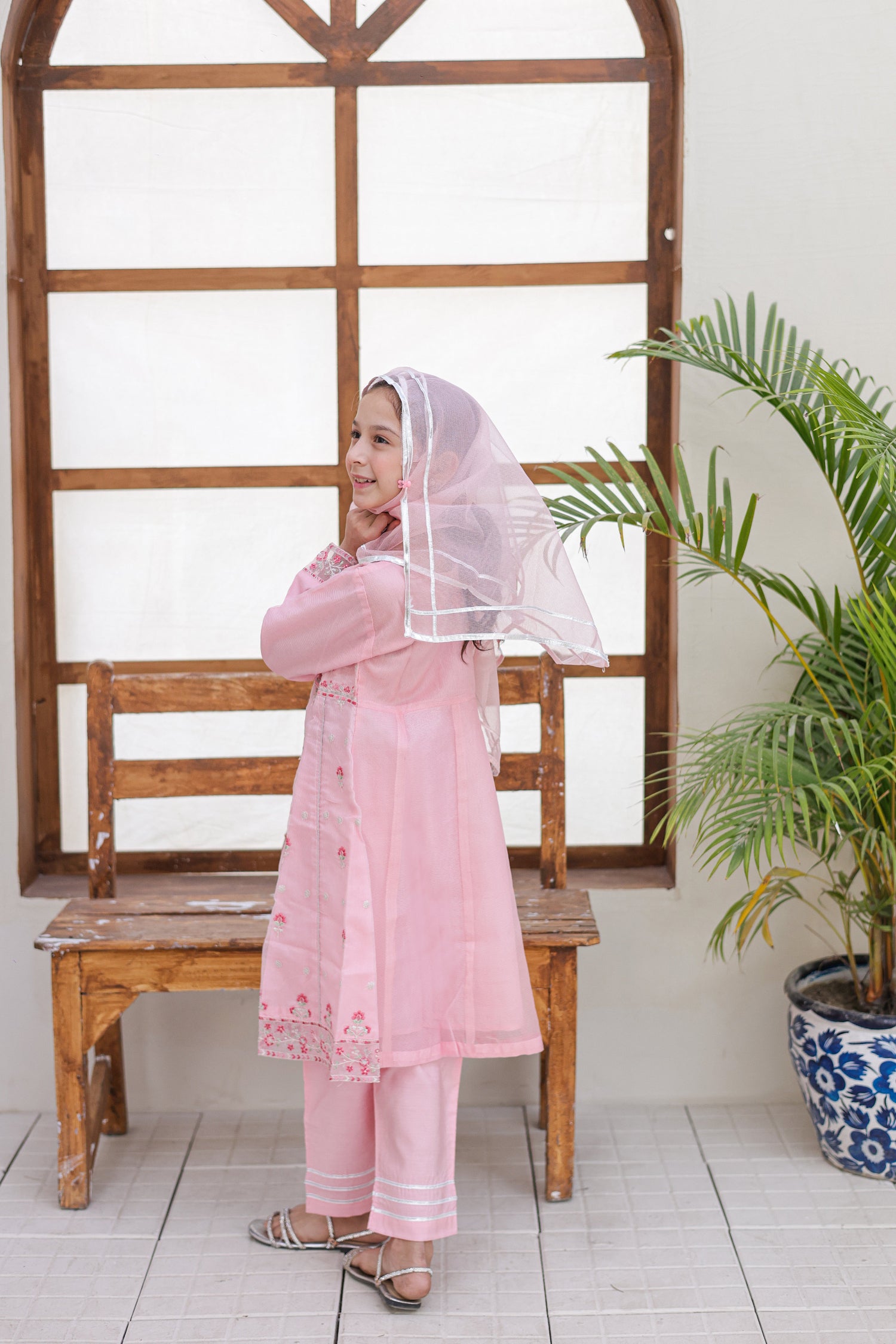 Child in a pink traditional outfit standing in a room with a wooden bench and plant.