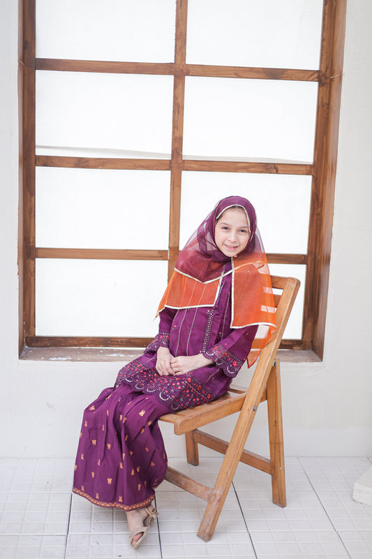 Woman in a purple traditional outfit with an orange shawl sitting on a wooden chair in front of a window.