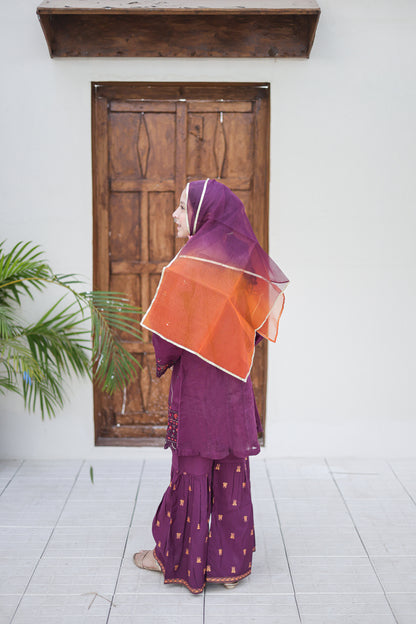 Woman in a purple outfit with an orange scarf standing in front of a wooden door.