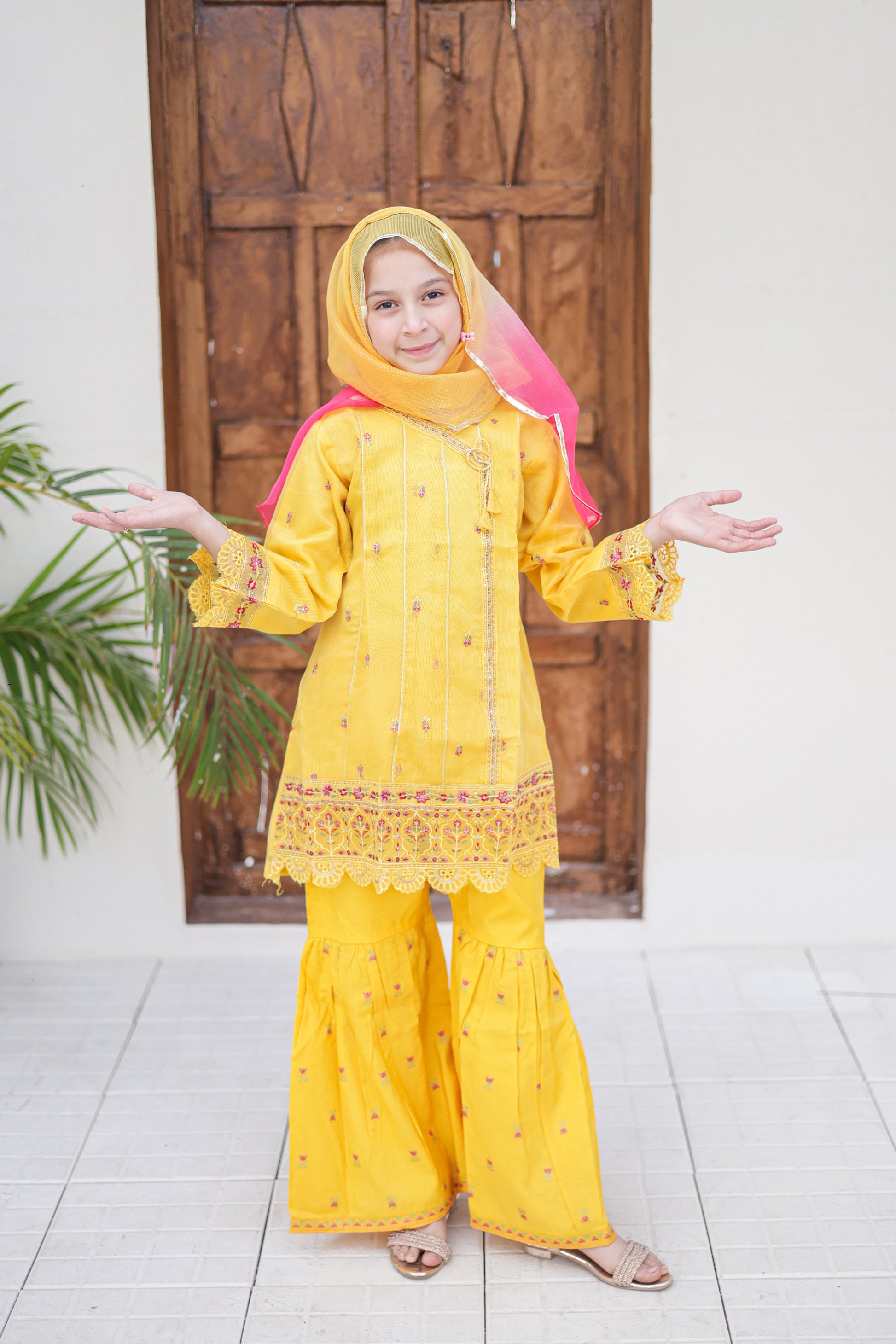 Child wearing a yellow traditional outfit with a pink hood in front of a wooden door.