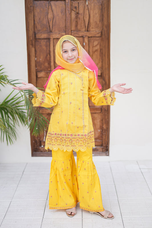 Child wearing a yellow traditional outfit with a pink hood in front of a wooden door.
