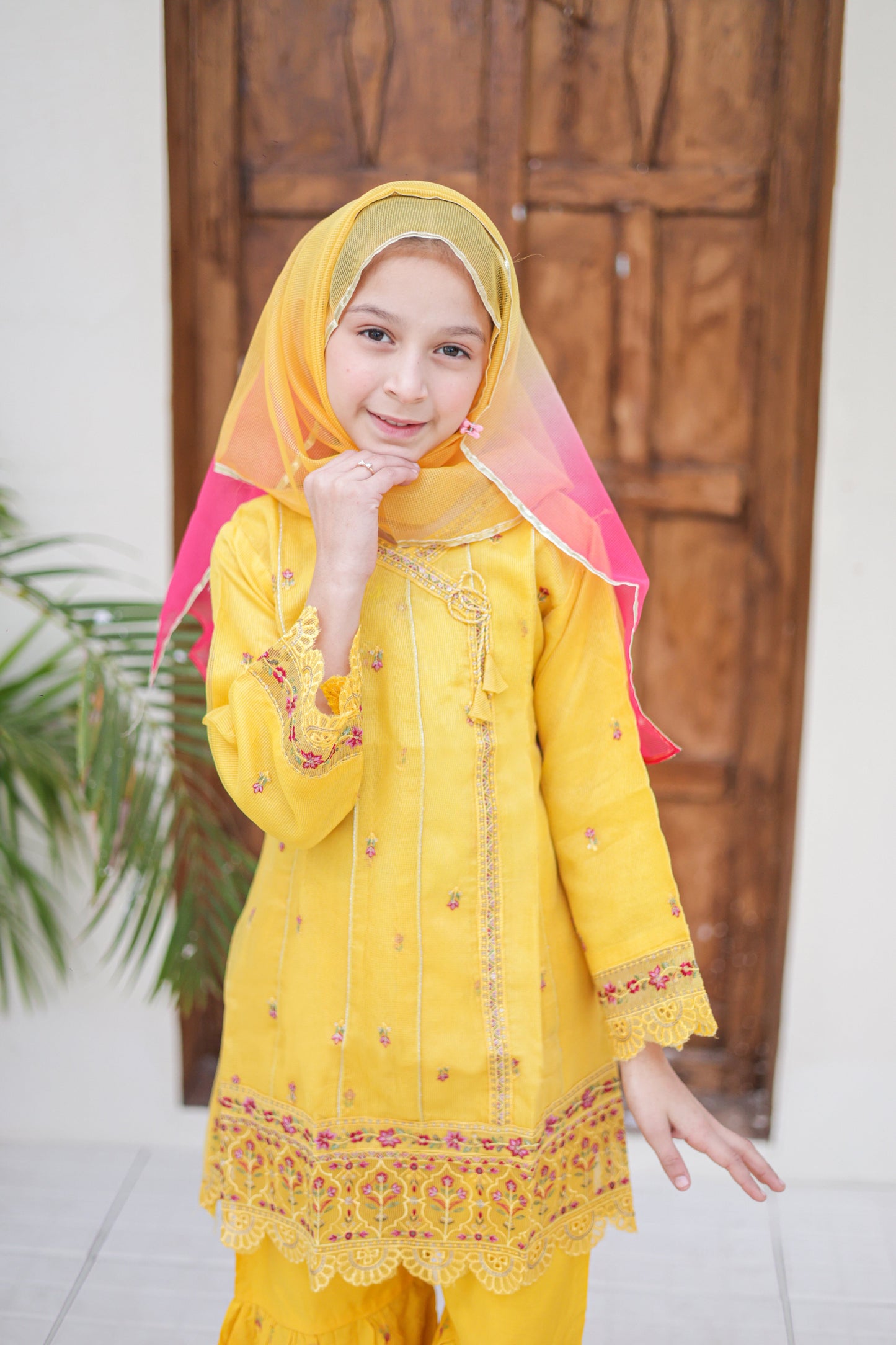 Young girl in a yellow traditional outfit with a pink dupatta standing in front of a wooden door.