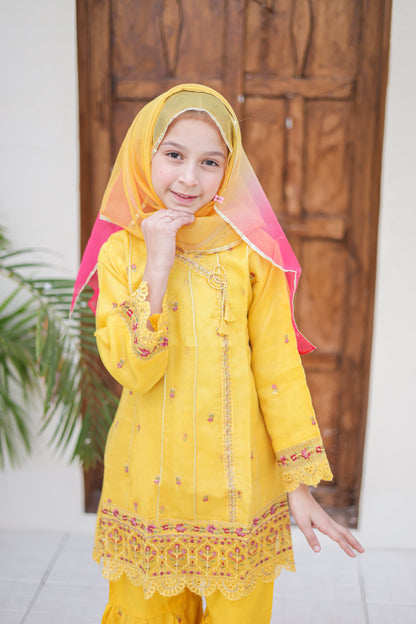 Young girl in a yellow traditional outfit with a pink dupatta standing in front of a wooden door.