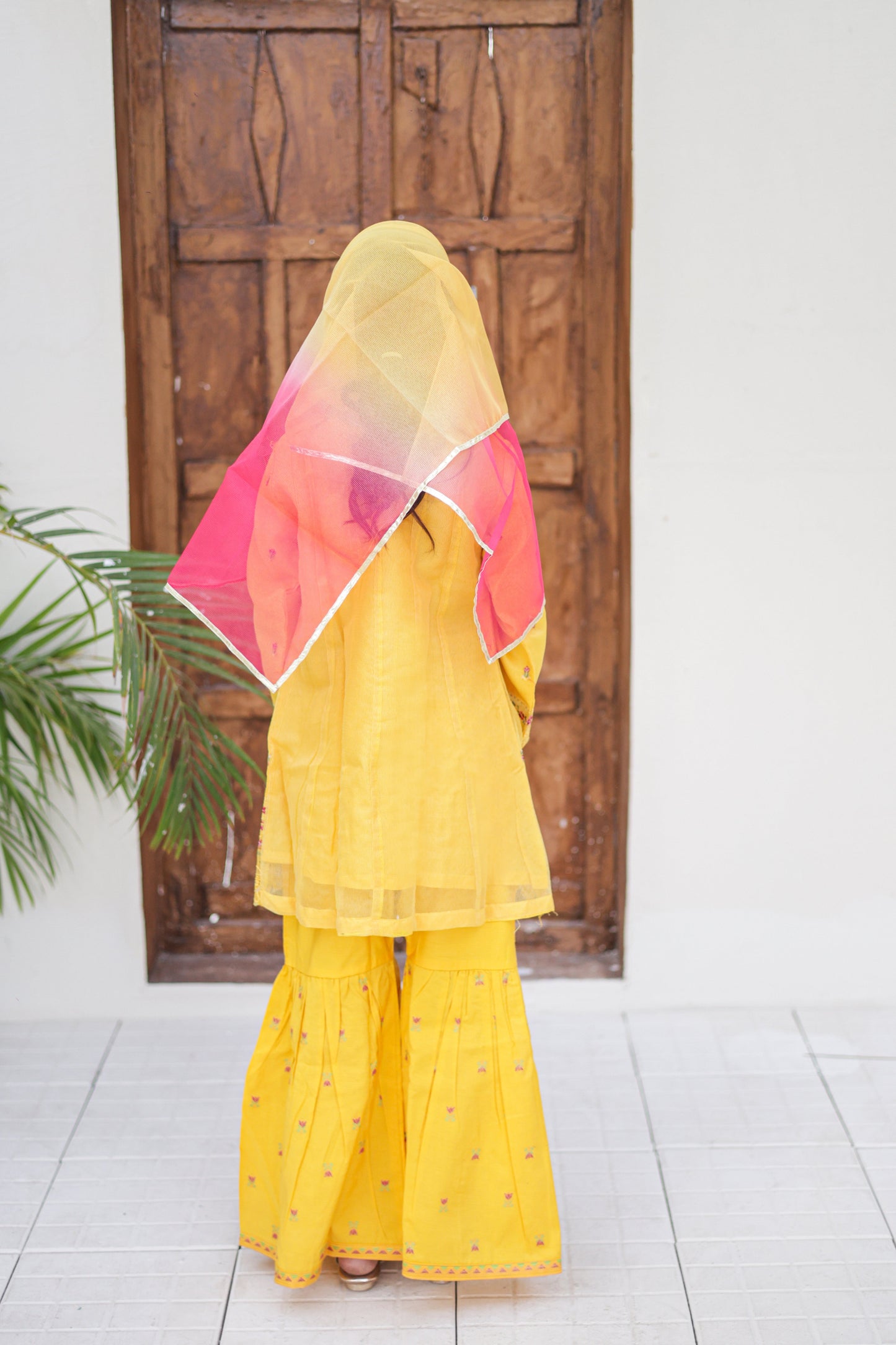 Person wearing a yellow outfit with a colorful headscarf in front of a wooden door.