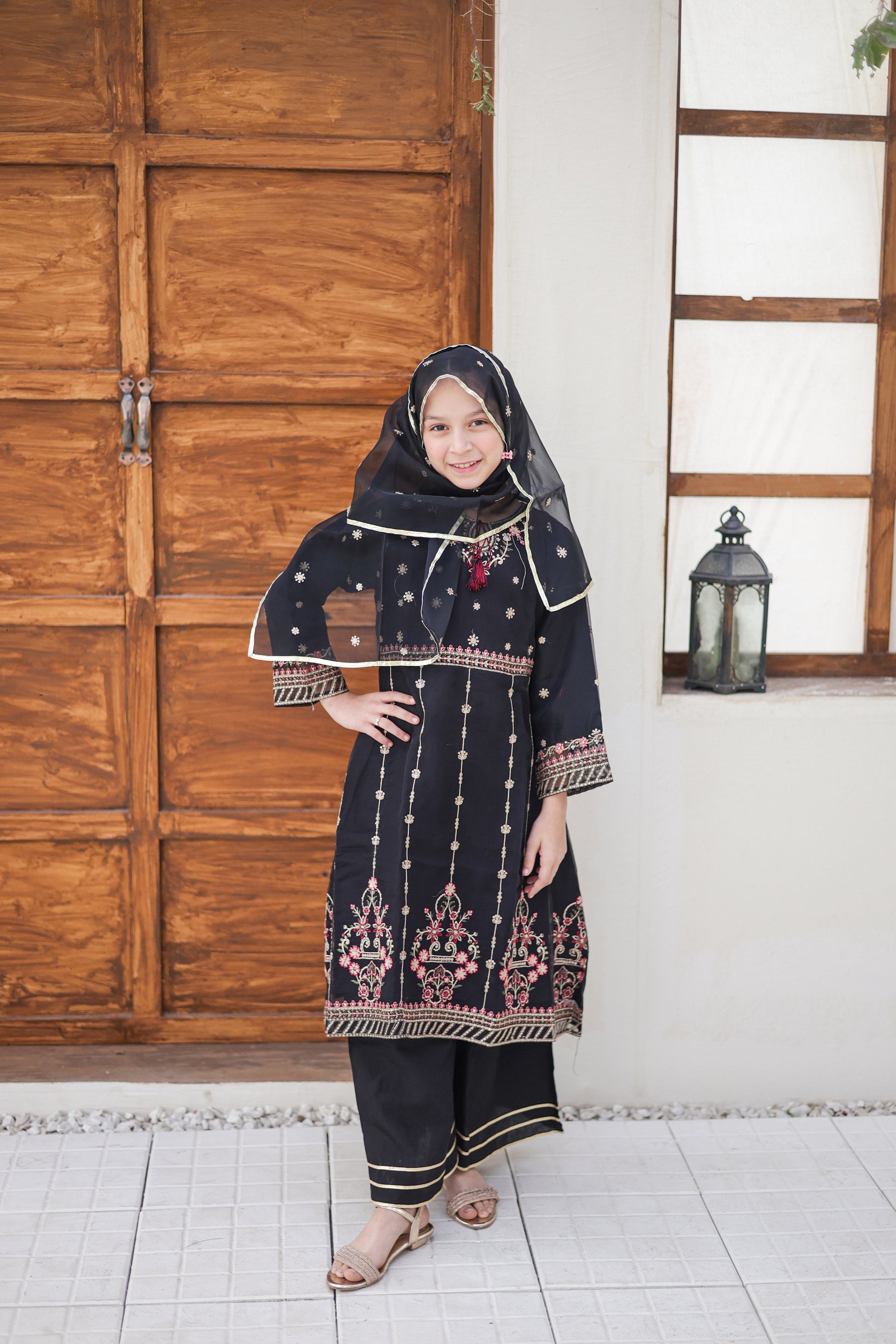 Woman wearing a traditional outfit with intricate patterns in a room with wooden doors and a window.