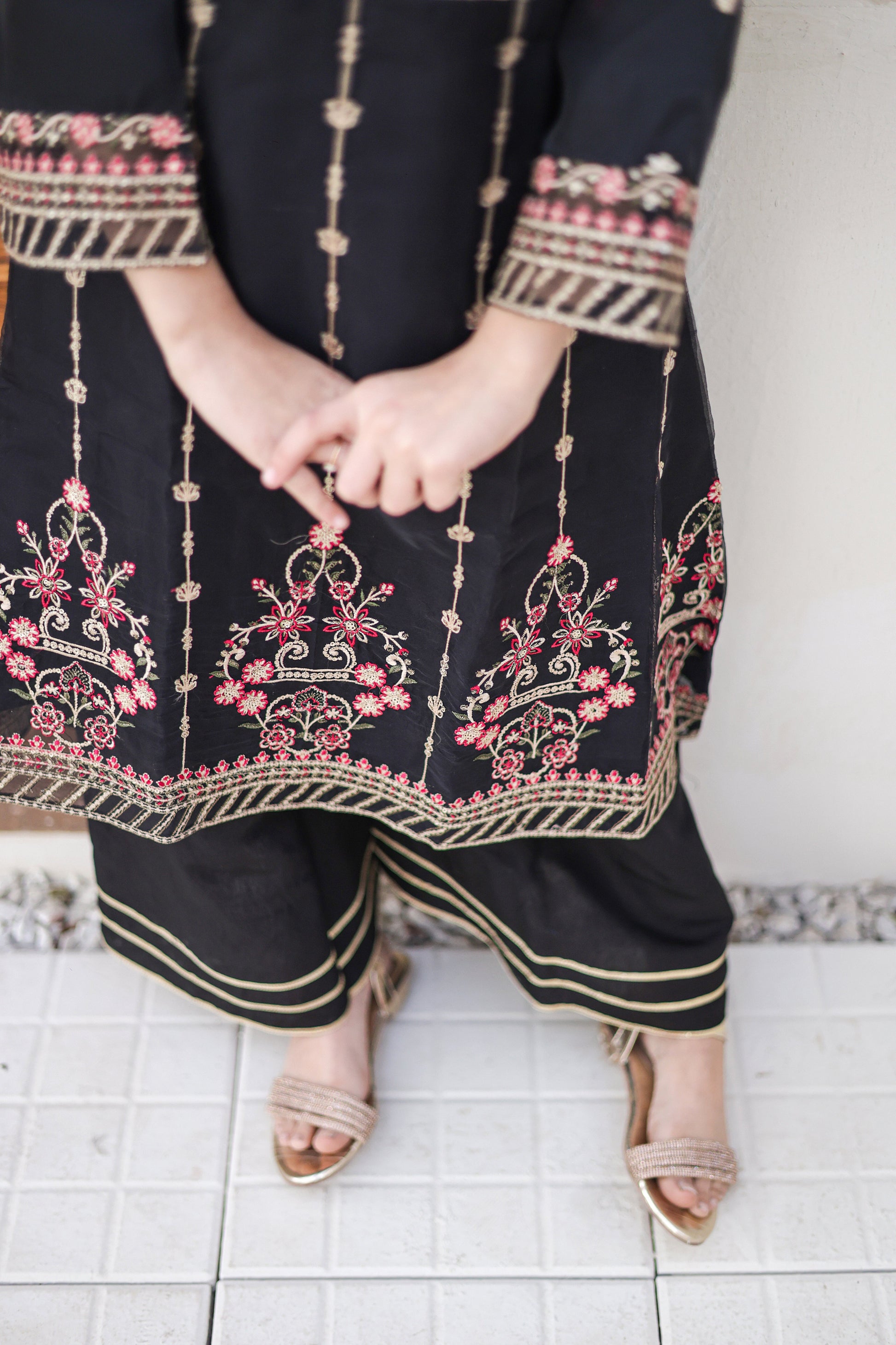 Person wearing a black outfit with intricate pink and gold embroidery on a tiled floor.
