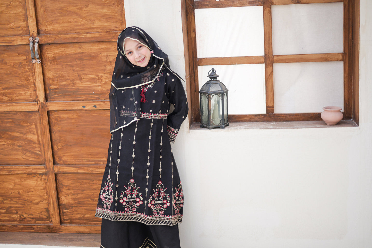 Woman in traditional black embroidered dress standing in a room with wooden paneling and a window.