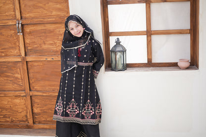 Woman in traditional black embroidered dress standing in a room with wooden paneling and a window.