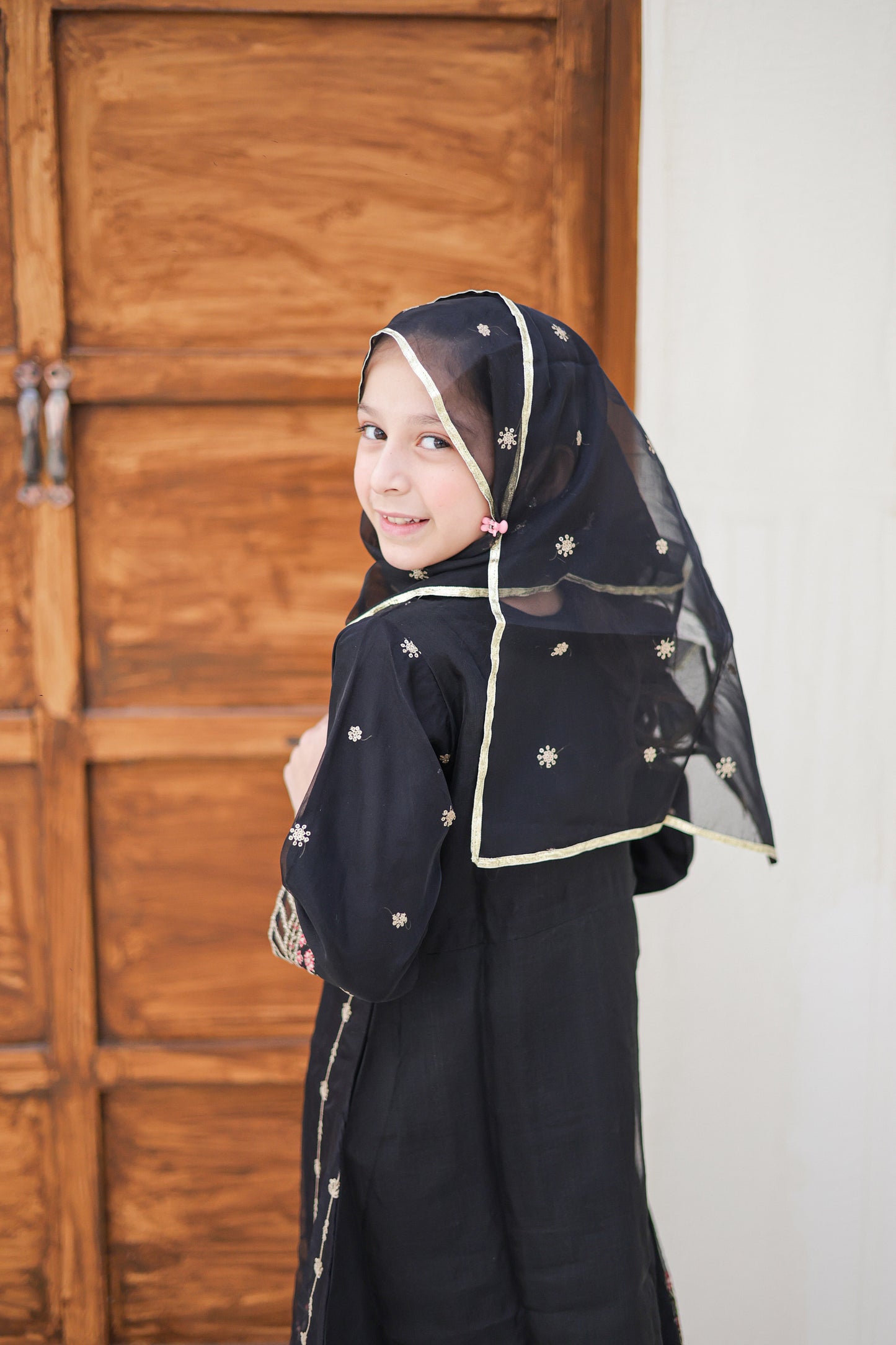 Young girl wearing a black abaya with a headscarf in front of a wooden door.