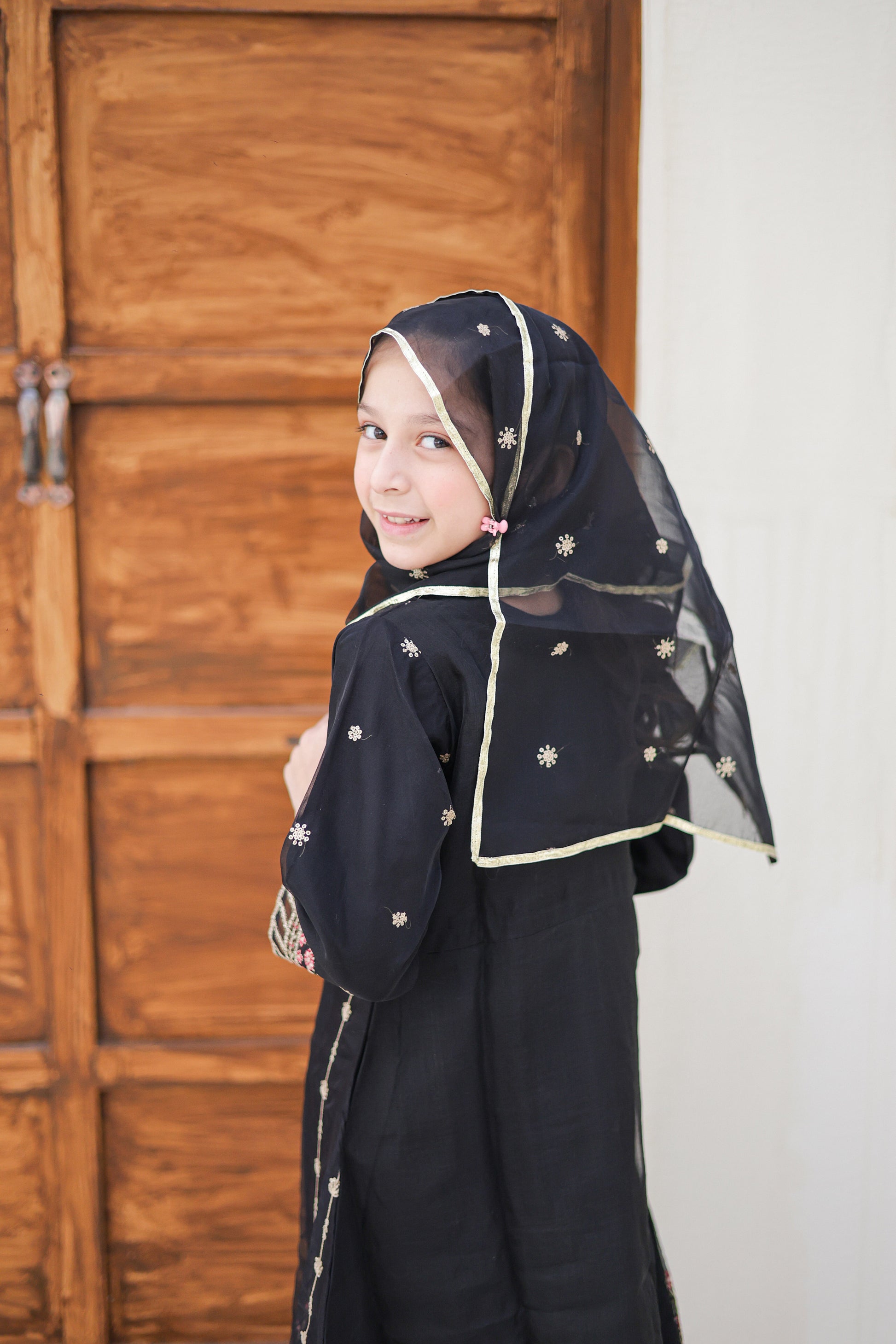 Young girl wearing a black abaya with a headscarf in front of a wooden door.