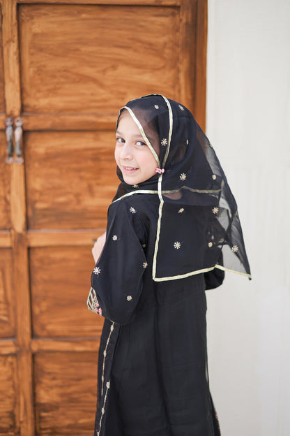 Young girl wearing a black abaya with a headscarf in front of a wooden door.