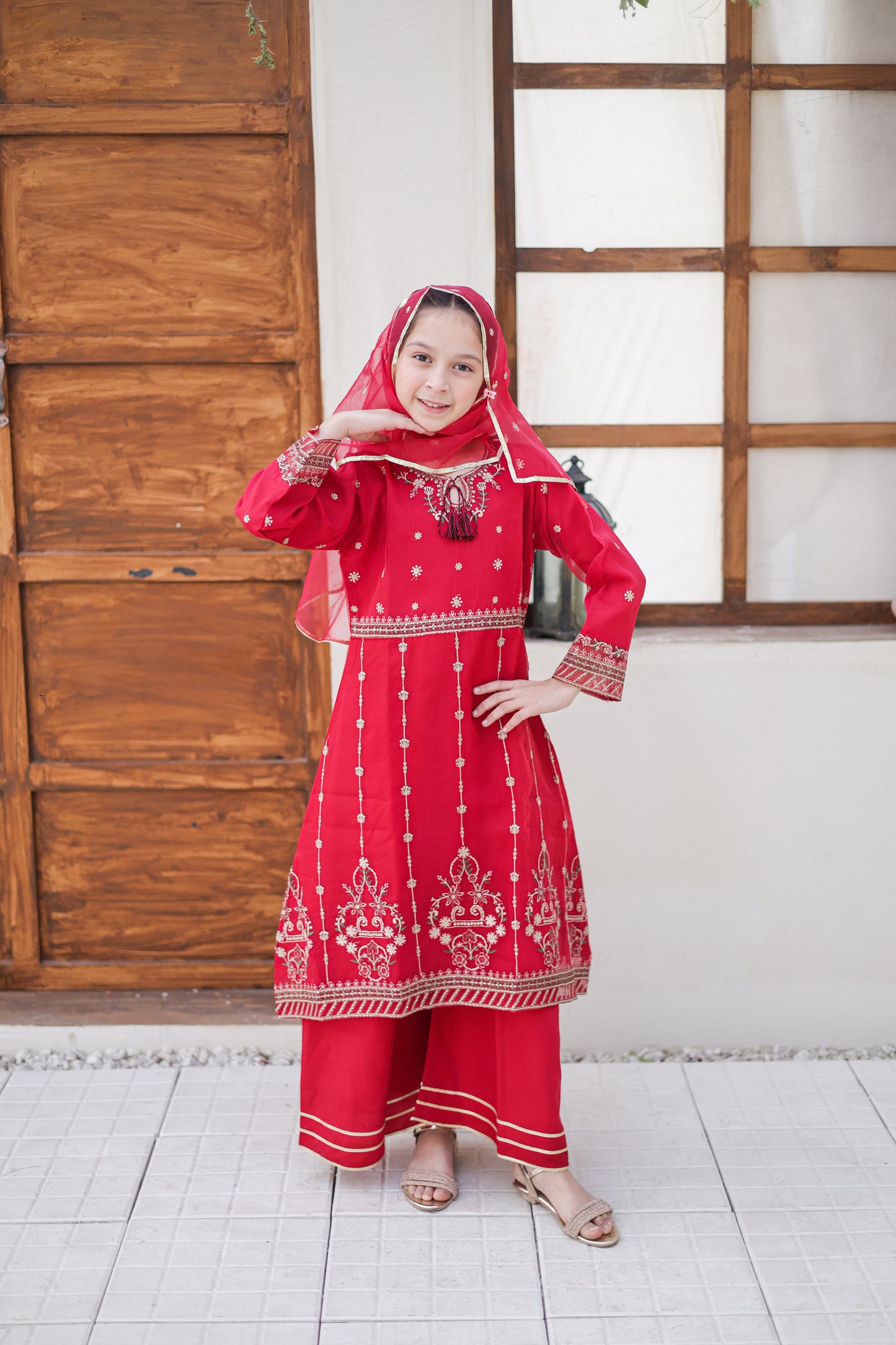 Person wearing a red traditional outfit with white patterns in a room with wooden doors and a white wall.