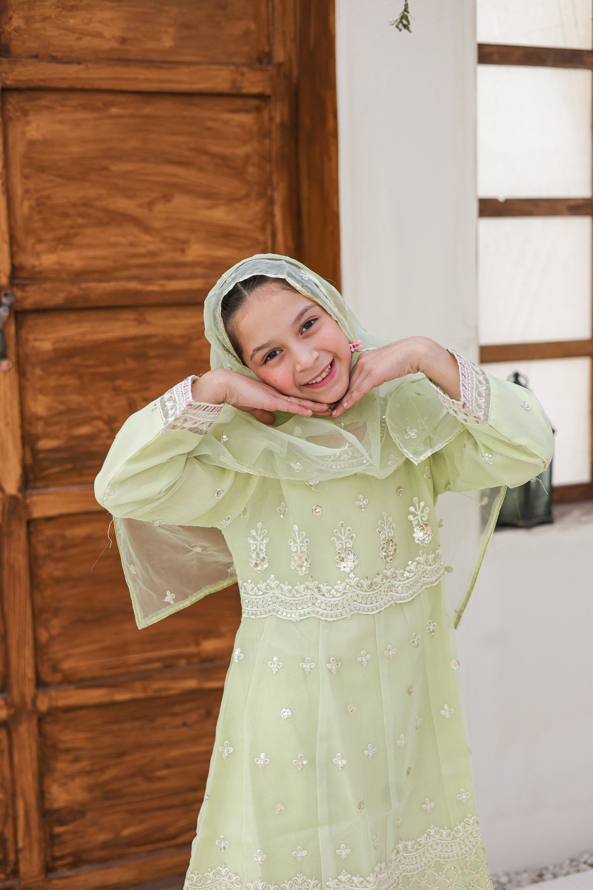 Young girl in a light green traditional outfit with floral embroidery, smiling indoors.