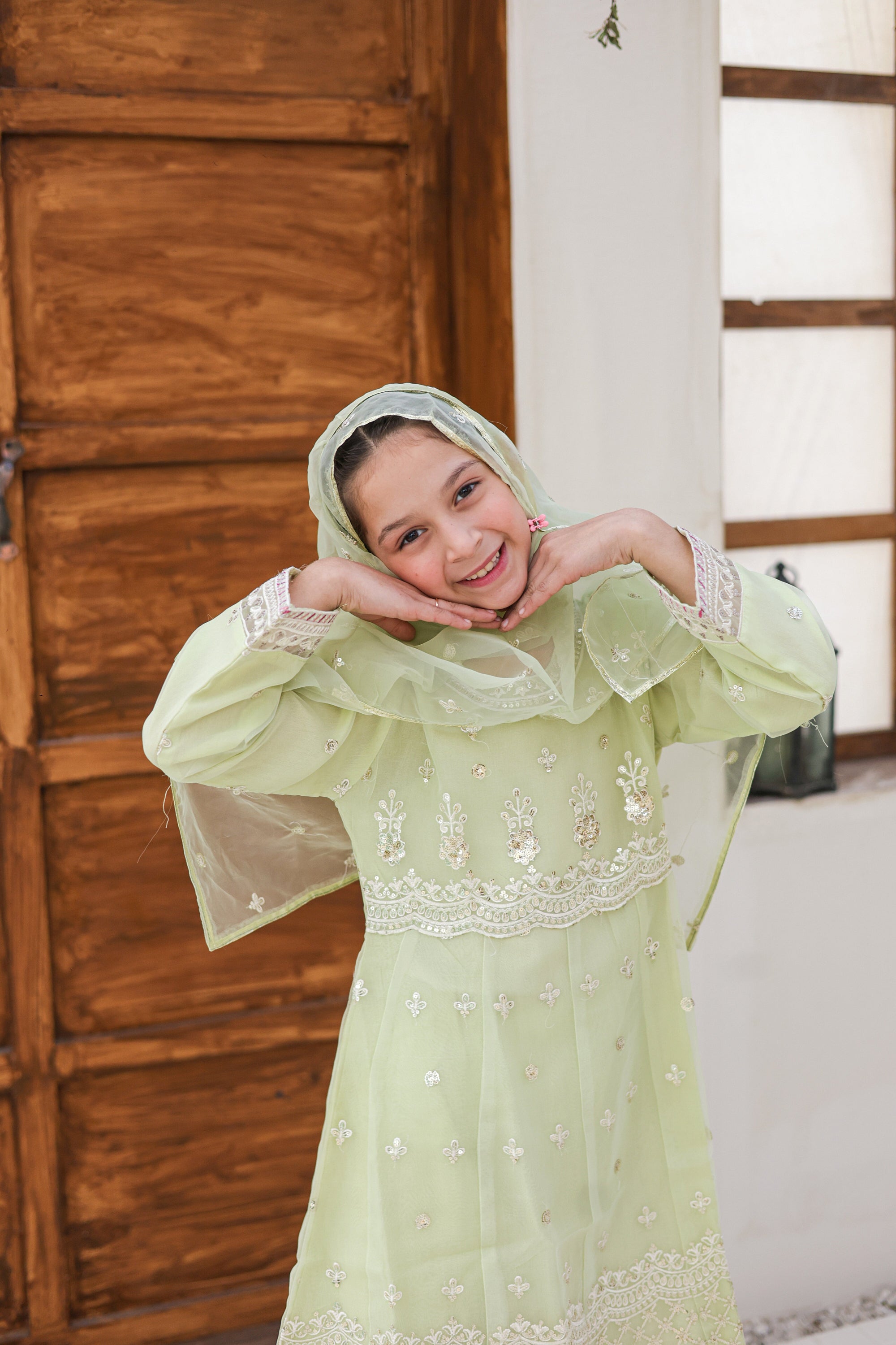 Young girl in a light green traditional outfit with floral embroidery, smiling indoors.