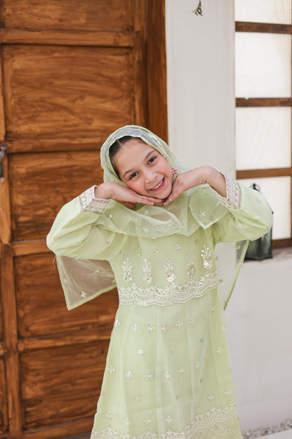 Young girl in a light green traditional outfit with floral embroidery, smiling indoors.