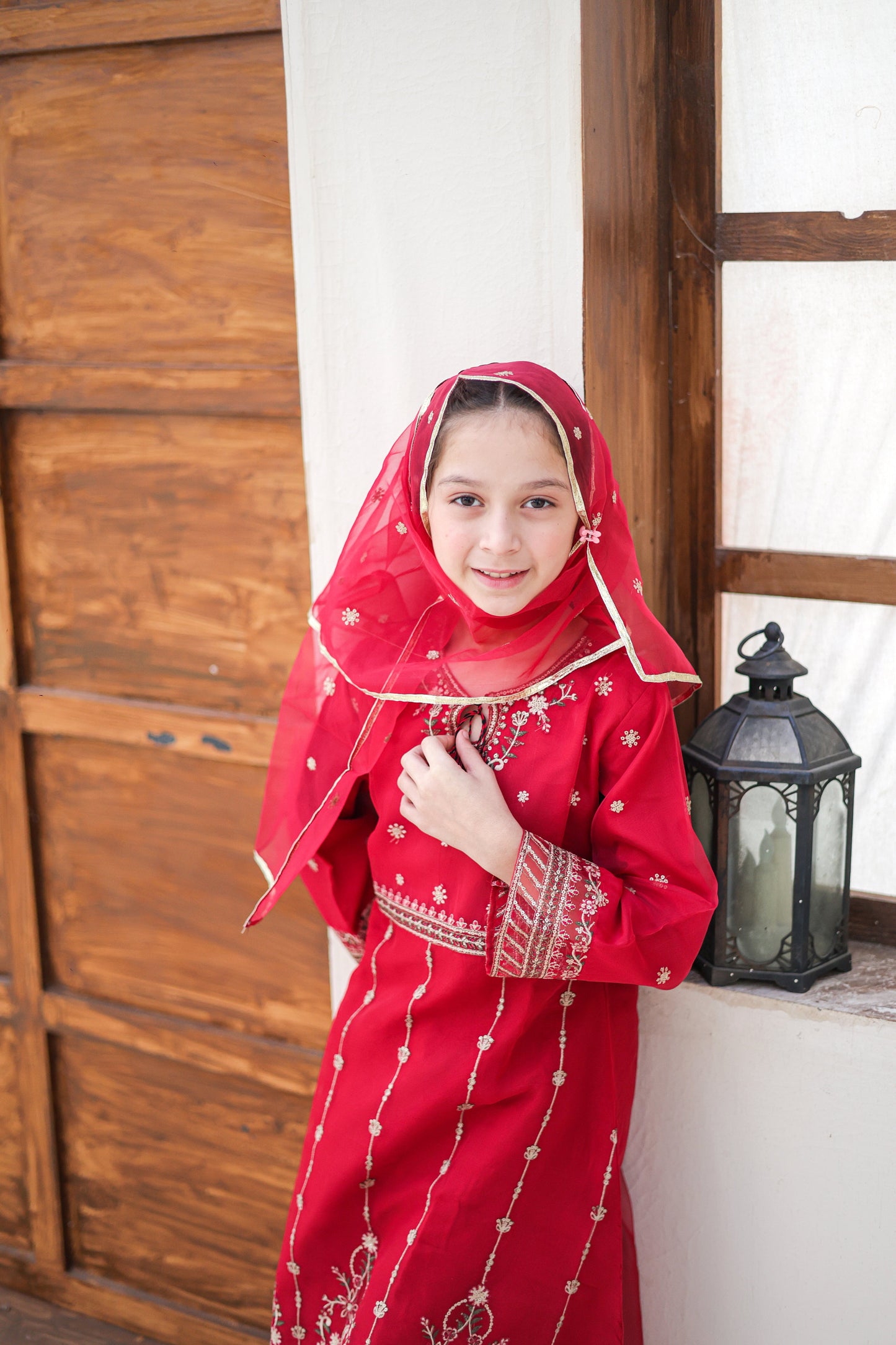 Young girl in a red traditional outfit with a matching headscarf, standing indoors.