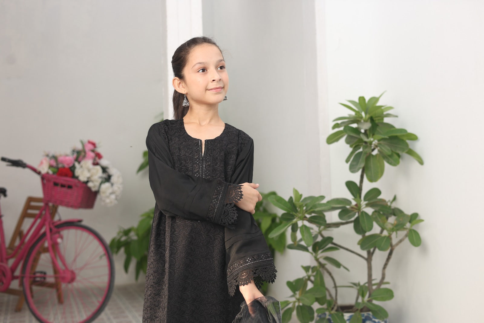 Young girl in a black dress standing indoors with plants and a pink bicycle in the background