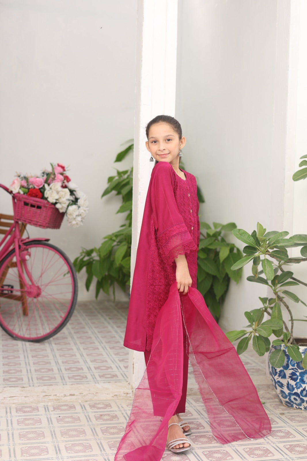 Young girl in a pink traditional outfit standing indoors with a bicycle and plants in the background.