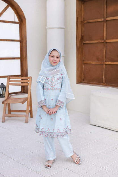 Young girl in a light blue traditional outfit standing indoors with wooden furniture and white walls.