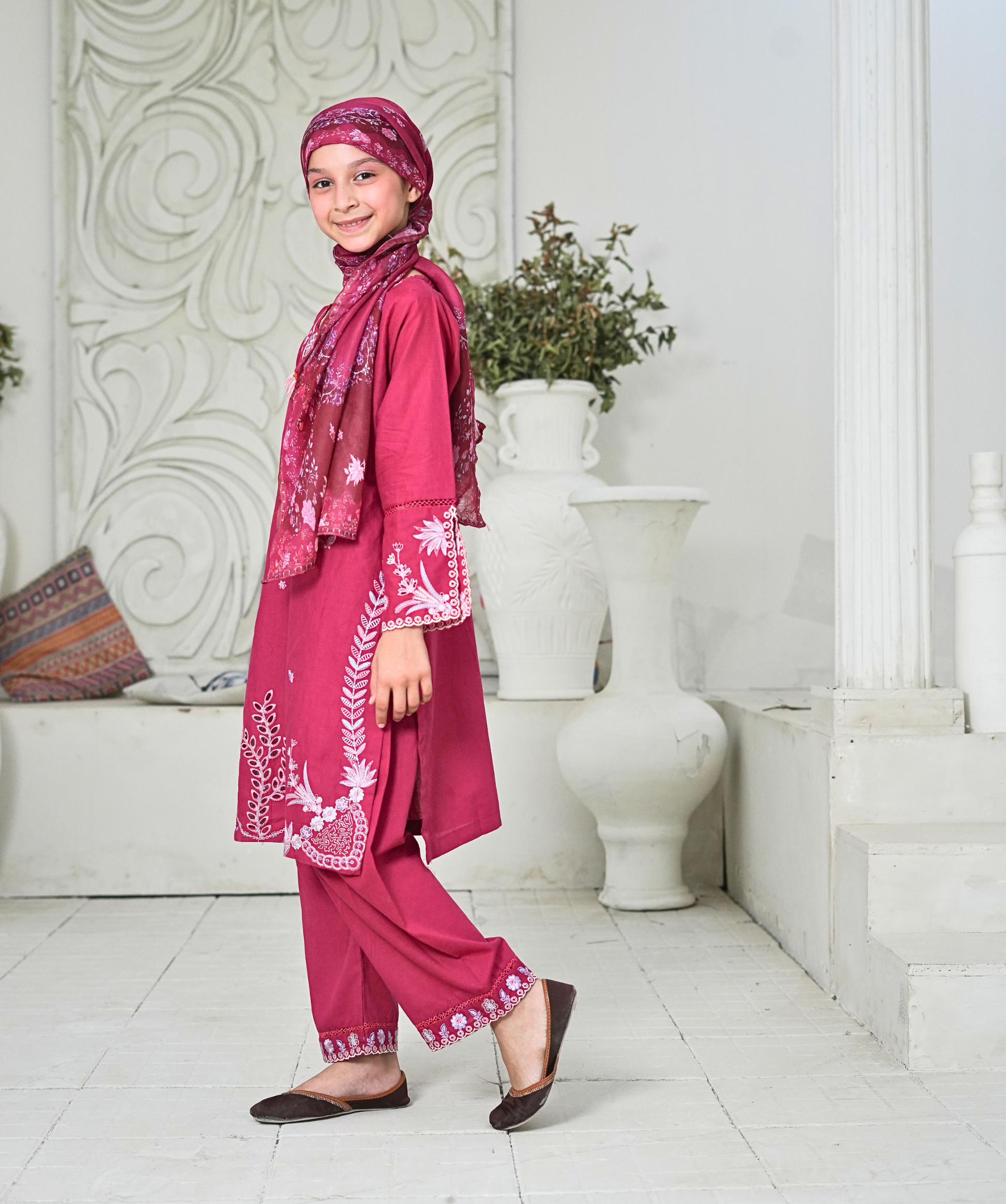 Young girl in a pink traditional outfit with a matching headscarf standing in a decorative indoor setting.