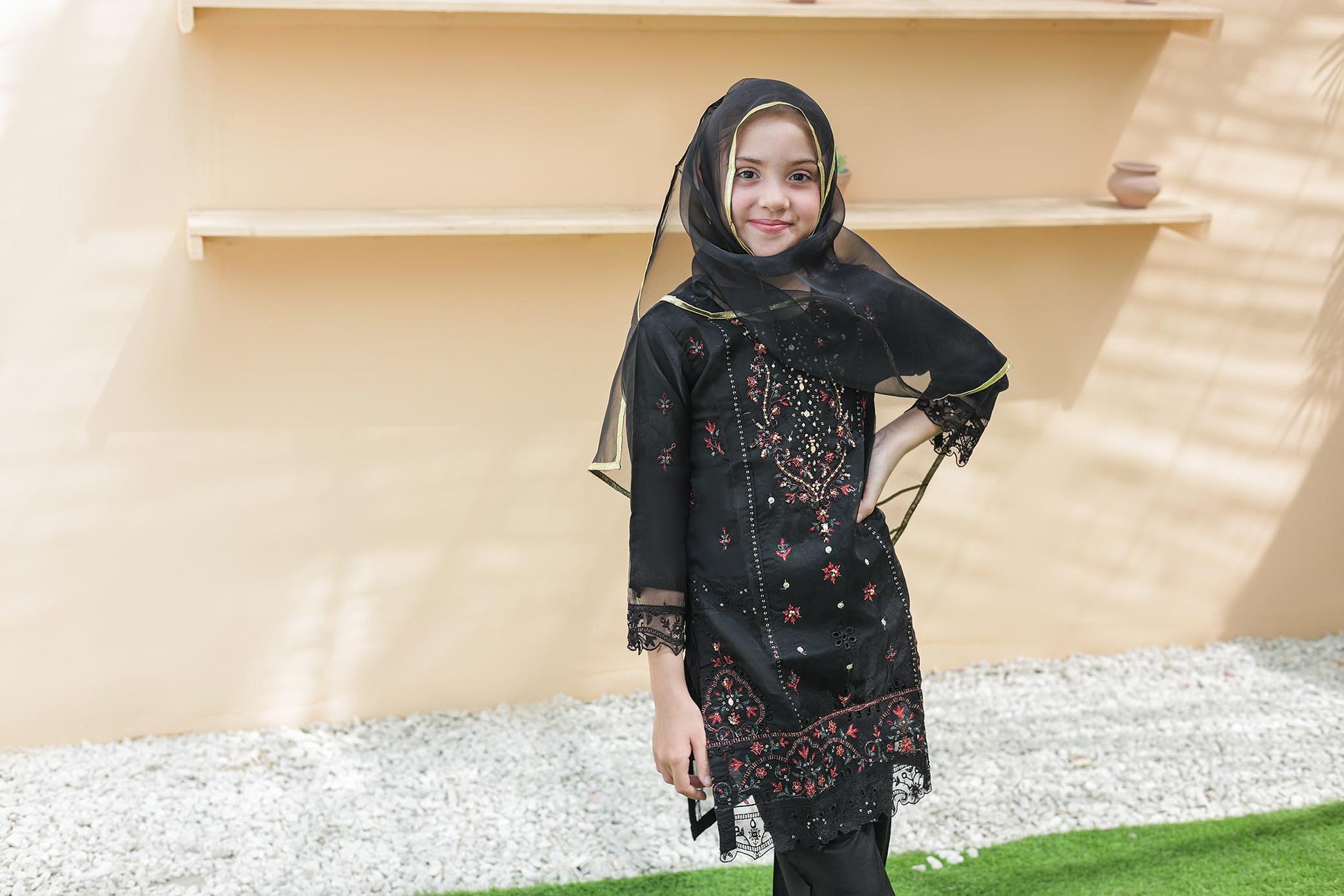 Child wearing a black embroidered outfit with a headscarf in an outdoor setting.