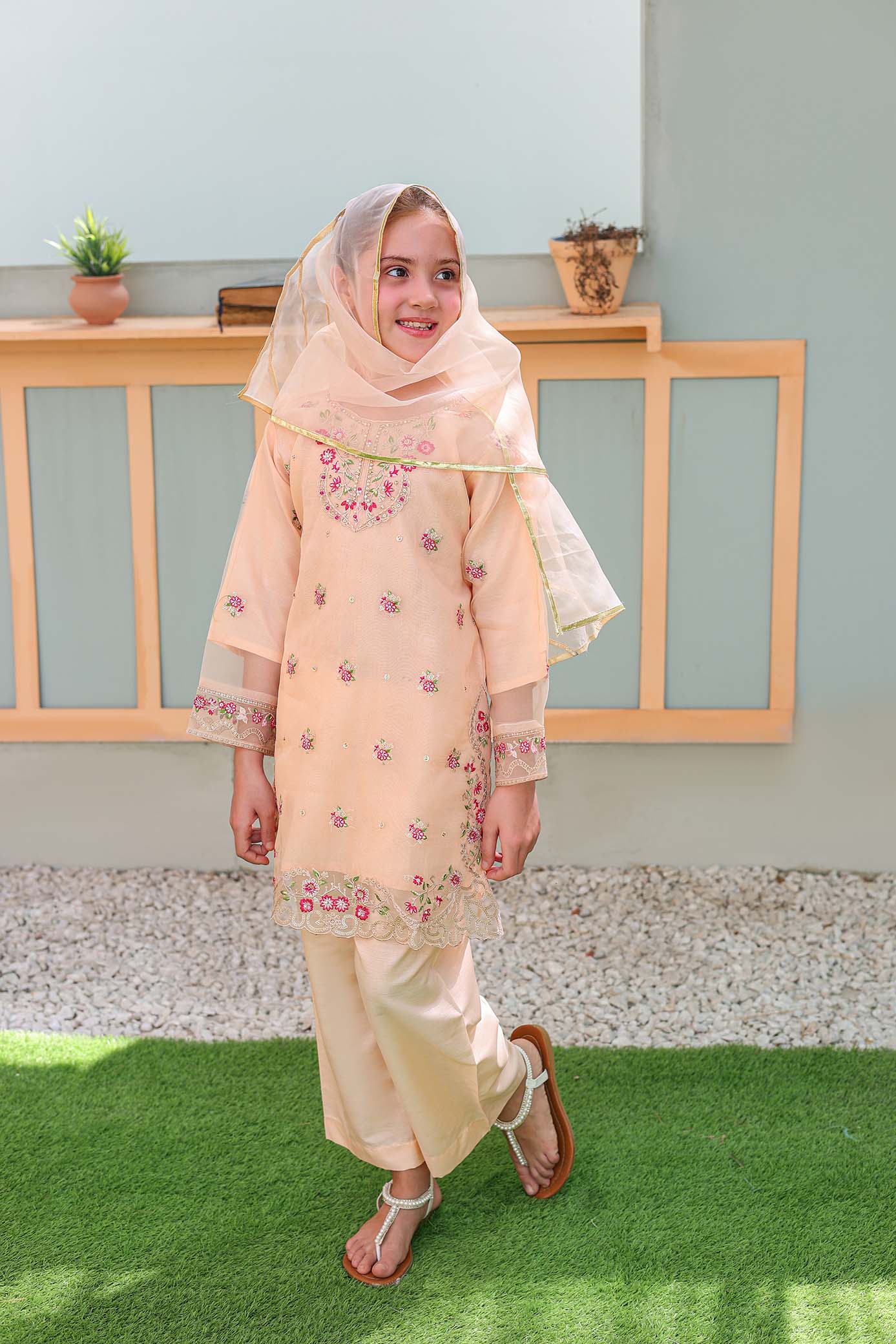 Young girl in a peach traditional outfit standing on grass with a light blue wall and wooden railing in the background.