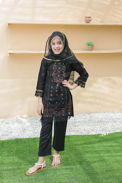 Woman in a black traditional outfit with floral patterns standing on grass in front of a beige wall.