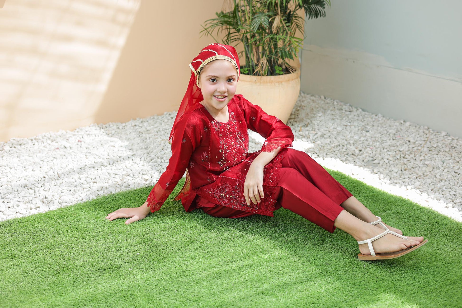 Child in a red outfit sitting on green grass with a plant in the background