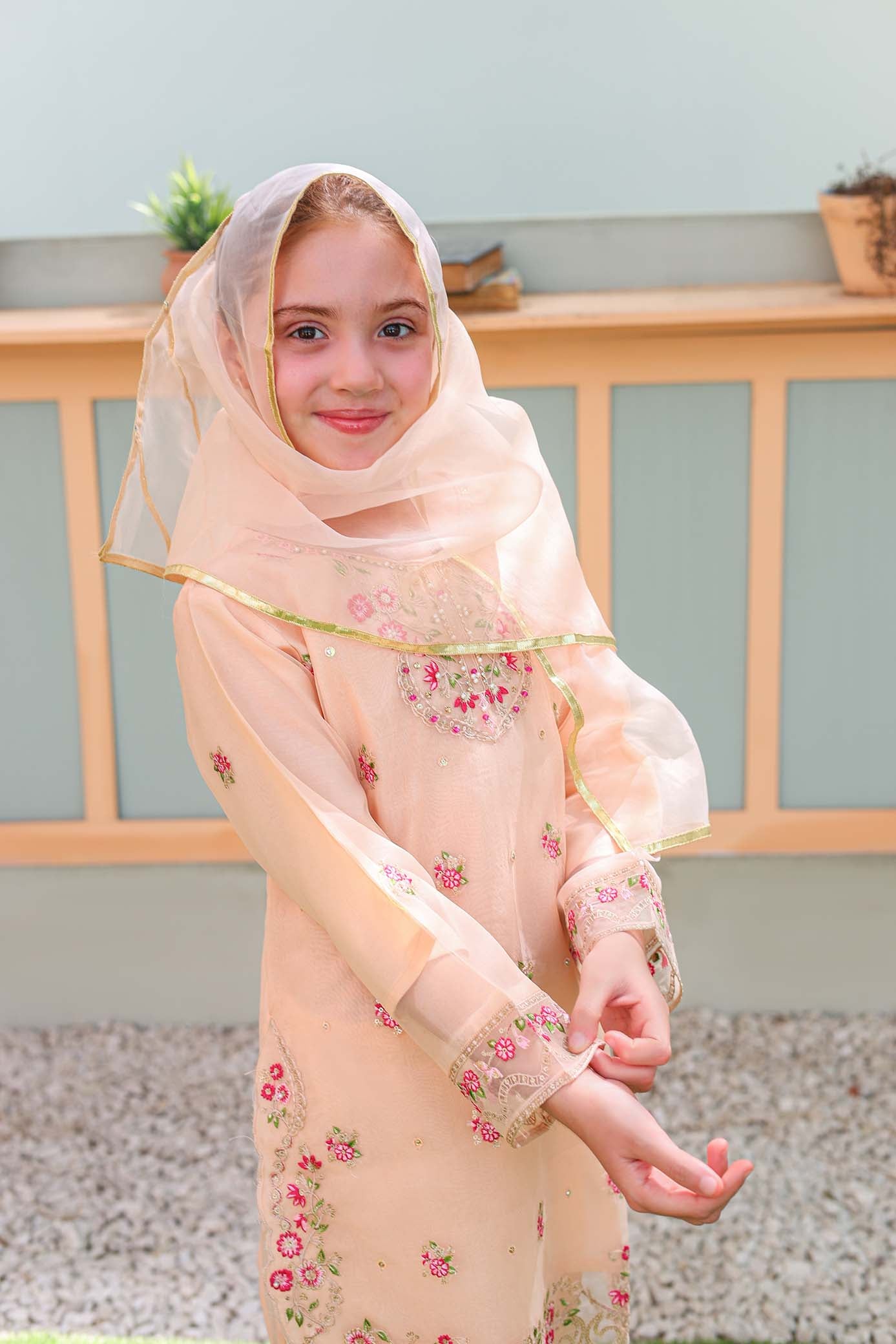 Young girl wearing a light pink floral dress and headscarf outdoors.