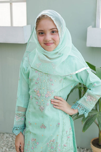Young girl wearing a light green embroidered dress and hijab outdoors.