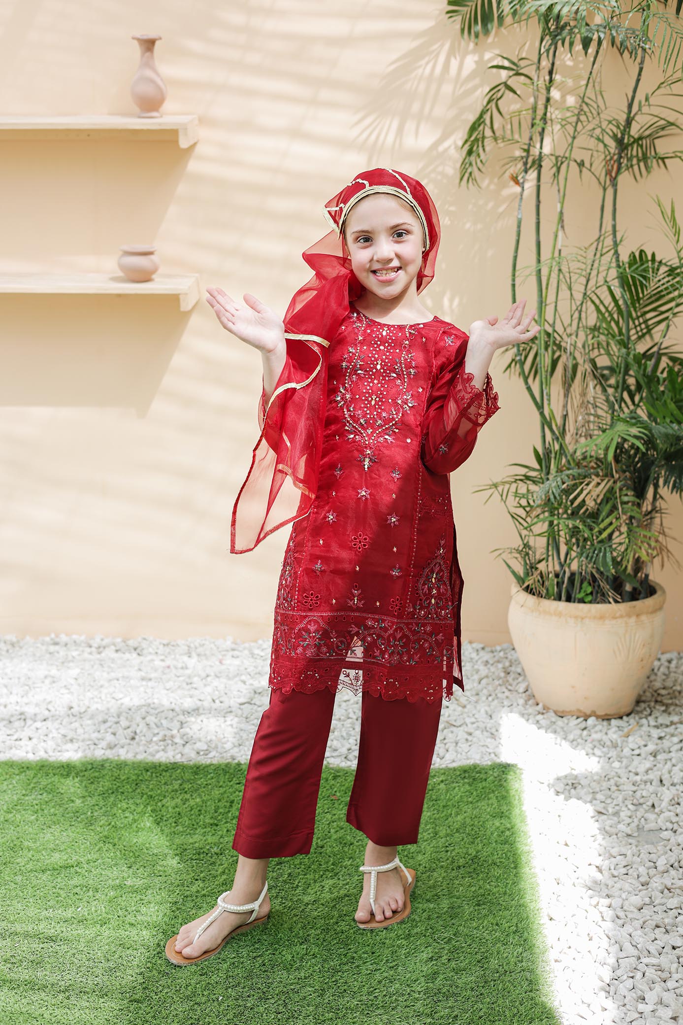 Child in a red traditional outfit standing outdoors with plants and a building in the background